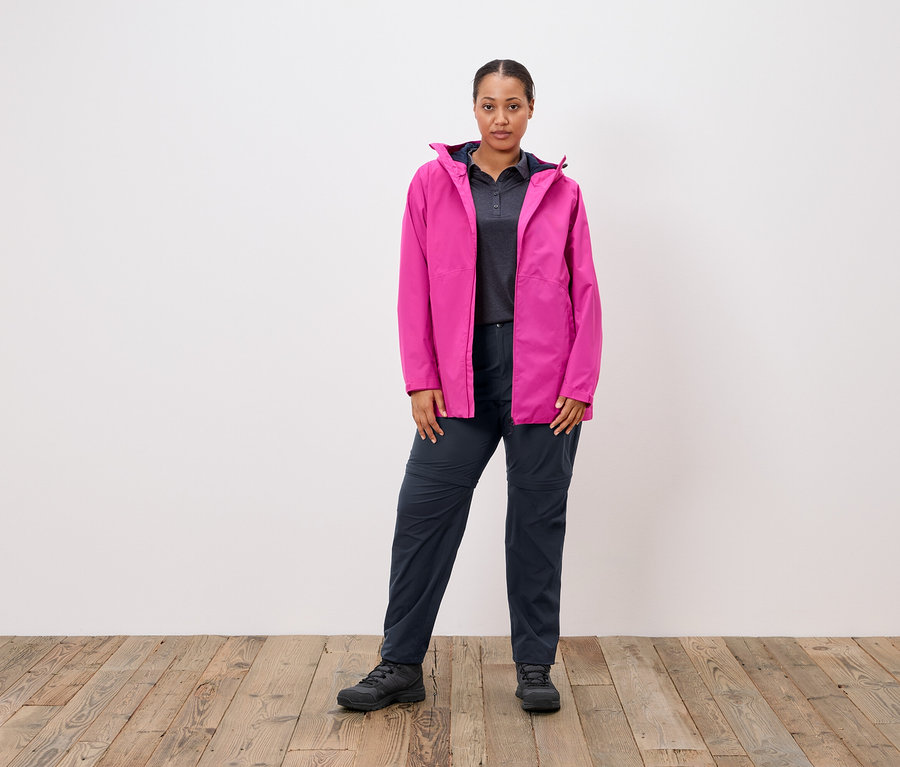 Žena stojí pred bielou stenou a má na sebe ružovú bundu, sivé polotričko a tmavomodré nohavice.