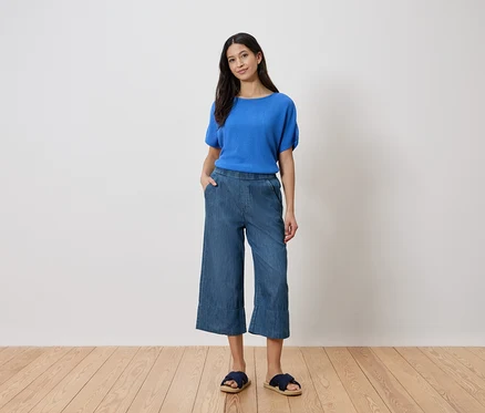 Žena stojí s rukou vo vrecku, má na sebe modrý top, džínsové culottes a sandále pred bielou stenou.