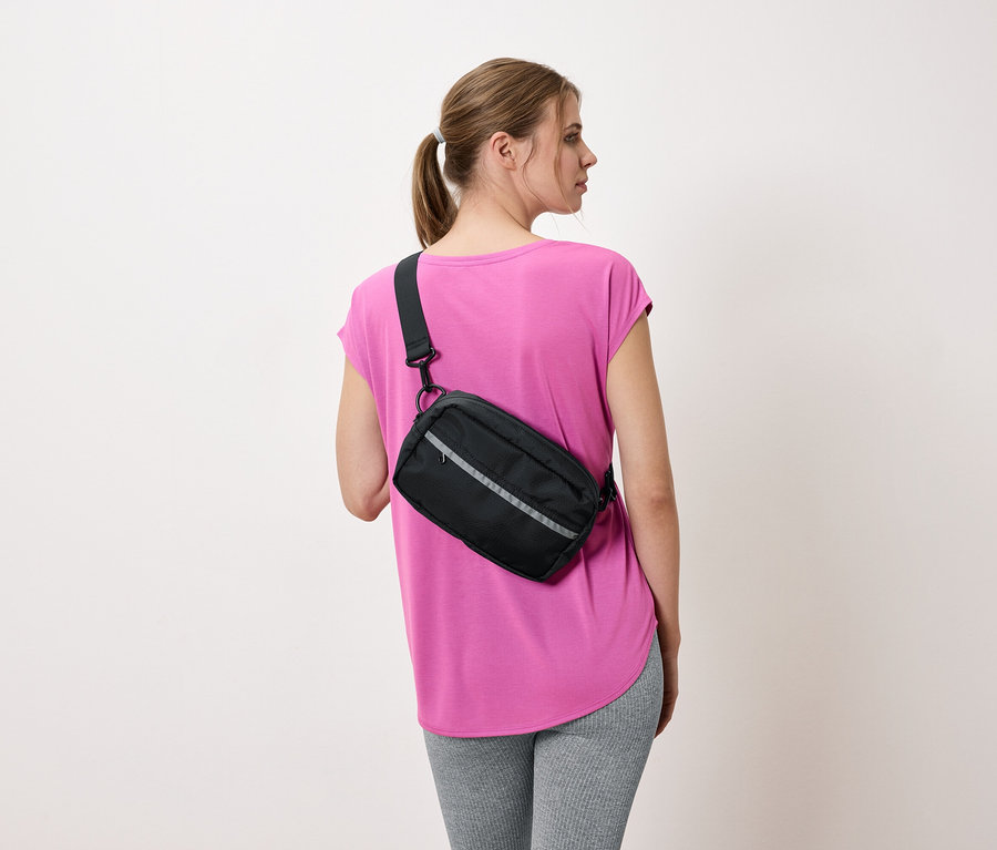 Žena zozadu s čiernou taškou cez plece, ružovým tričkom a sivými legínami.