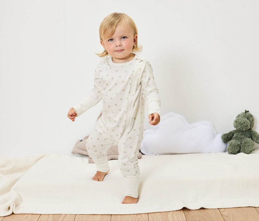 Blond dieťa v bielom overale s hviezdami stojí bosé na bielej deke vedľa obláčikového vankúša a zelenej plyšovej hračky.