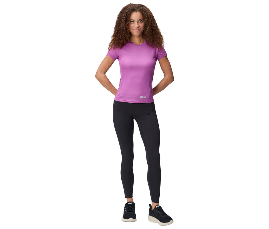 Žena stojí s rukami za chrbtom, má na sebe fialové tričko, čierne legíny a tenisky.