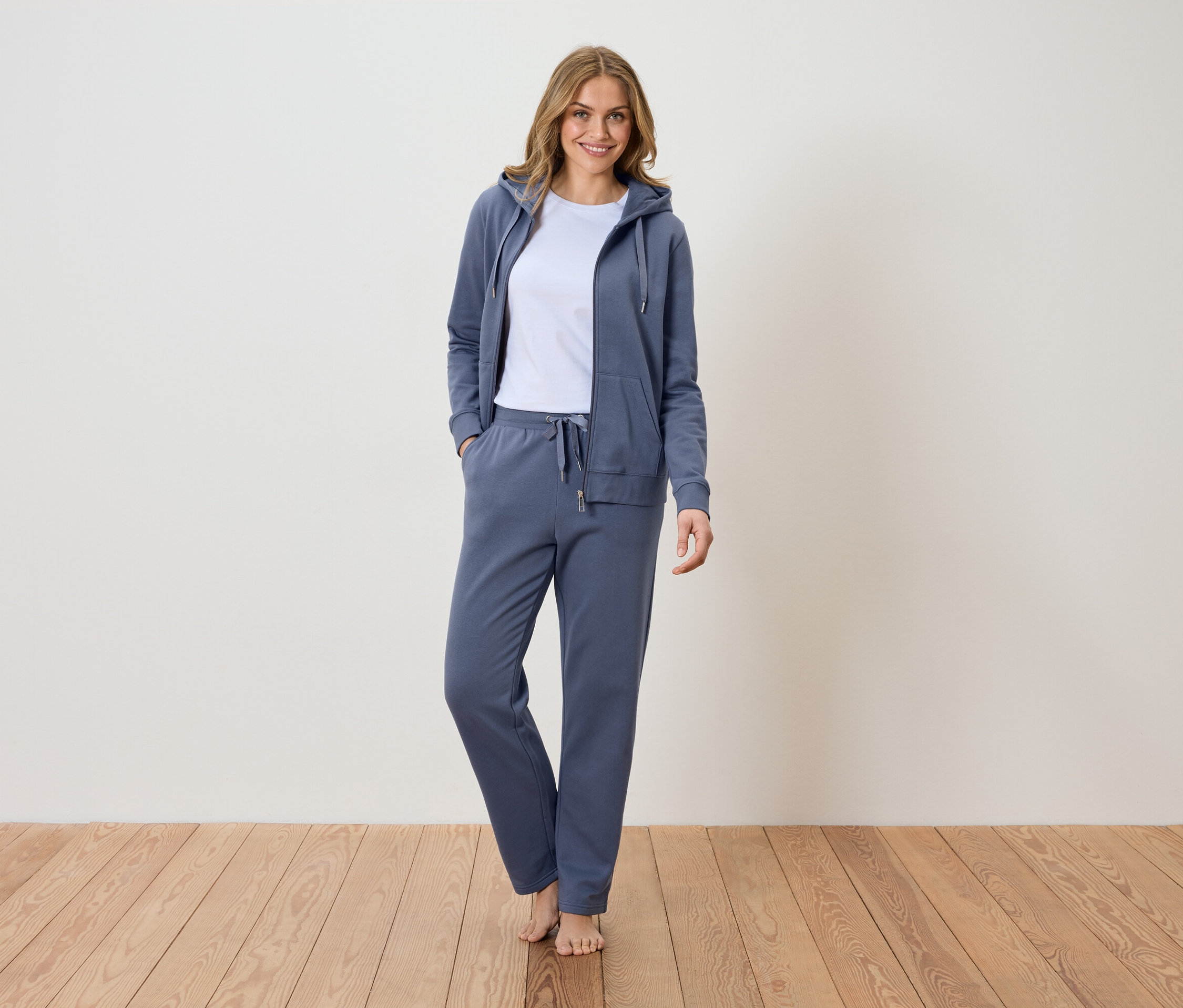 Žena stojí naboso na drevenej podlahe a má na sebe modrú teplákovú súpravu s mikinou s kapucňou a bielym tričkom.