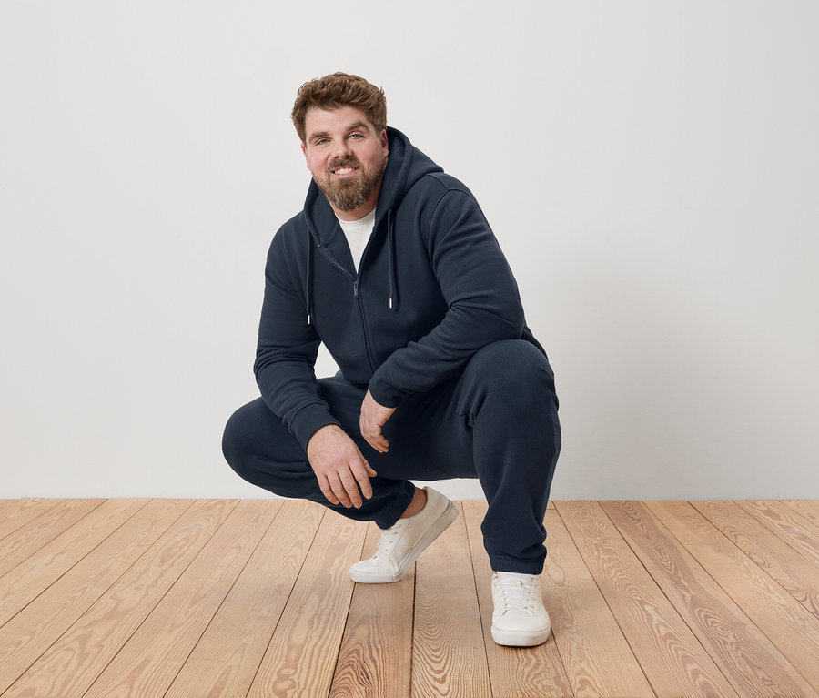 Muž čupí na drevenej podlahe, oblečený v tmavomodrej teplákovej súprave a bielych teniskách.