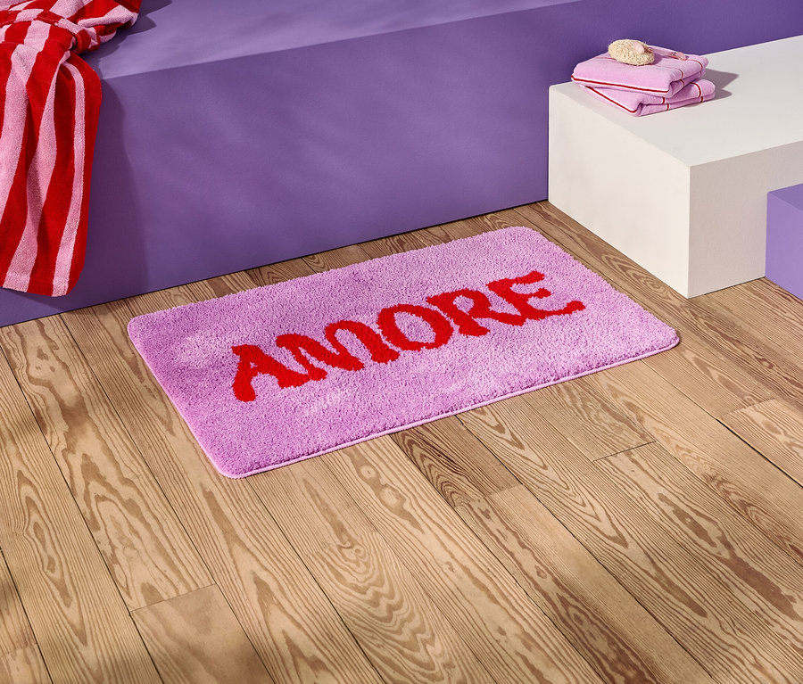 Na drevenej podlahe leží fialová kúpeľňová predložka s červeným nápisom "AMORE".