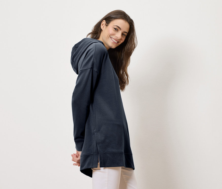 Žena má na sebe dlhú, tmavomodrú mikinu s kapucňou a svetlé nohavice, usmieva sa do kamery.