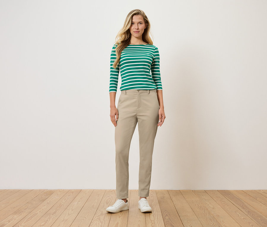 Žena stojí a má na sebe zeleno-biele pruhované tričko s dlhým rukávom, béžové nohavice a biele topánky.