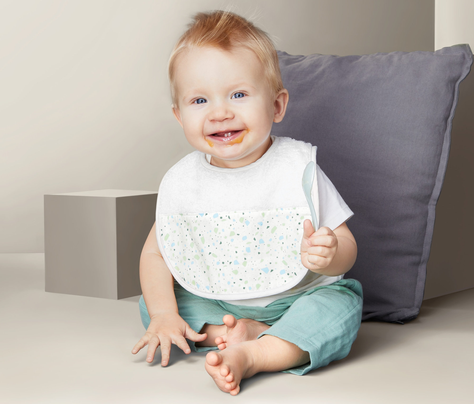 Usmievajúci sa dojča sedí na zemi s podbradníkom Detské podbradníky, 2 ks a lyžicou v ruke.