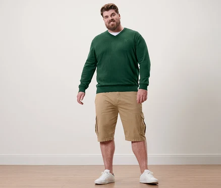 Muž stojí a má na sebe tmavozelený pulóver s výstrihom do V, khaki šortky a biele tenisky.