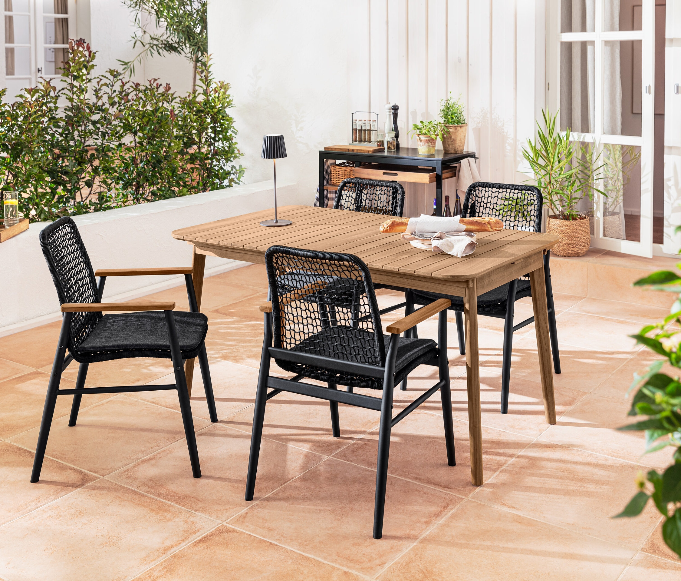 Štyri jedálenské stoličky s tíkovými opierkami rúk a textilným výpletom stoja okolo rozkladacieho stola z tíkového dreva. Na stole je položená LED akumulátorová stolová lampa do exteriéru, čierna.