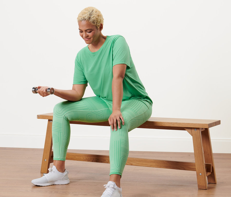 Žena sedí na lavici a cvičí s činkou v zelenom športovom tričku a obojstranných športových legínsoch.