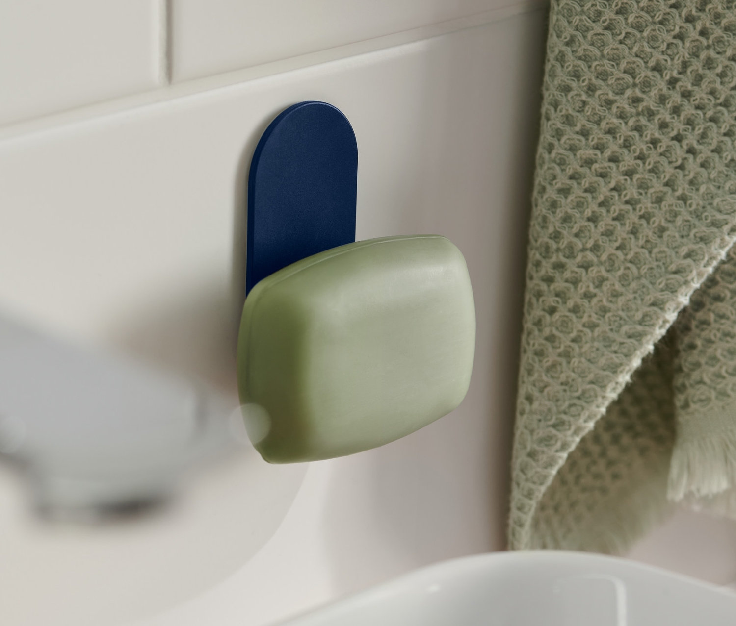 Zelené mydlo visí na magnetickom držiaku na mydlo modrej farby, ktorý je pripevnený na bielych kachličkách pri umývadle. Vpravo visí zelený uterák.