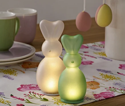 Dva LED zajačiky stoja na kvetinovom behúni, v pozadí šálky a veľkonočné vajíčka.