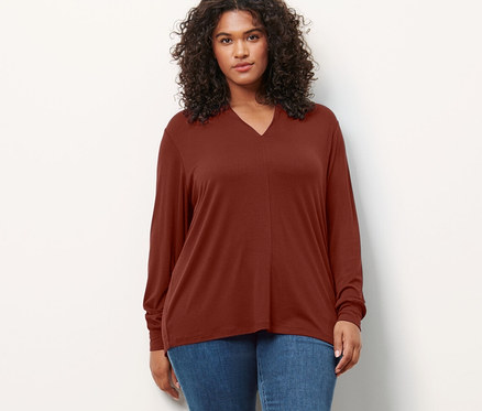 Žena stojí a má na sebe blúzkové tričko s výstrihom do V a tregínsy »Fit Nelly«.