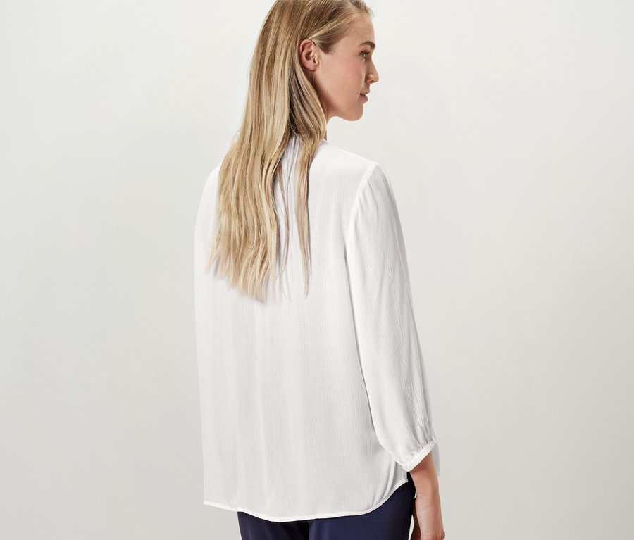 Blondínka pózuje chrbtom k fotoaparátu a má na sebe bielu tunikovú blúzku.