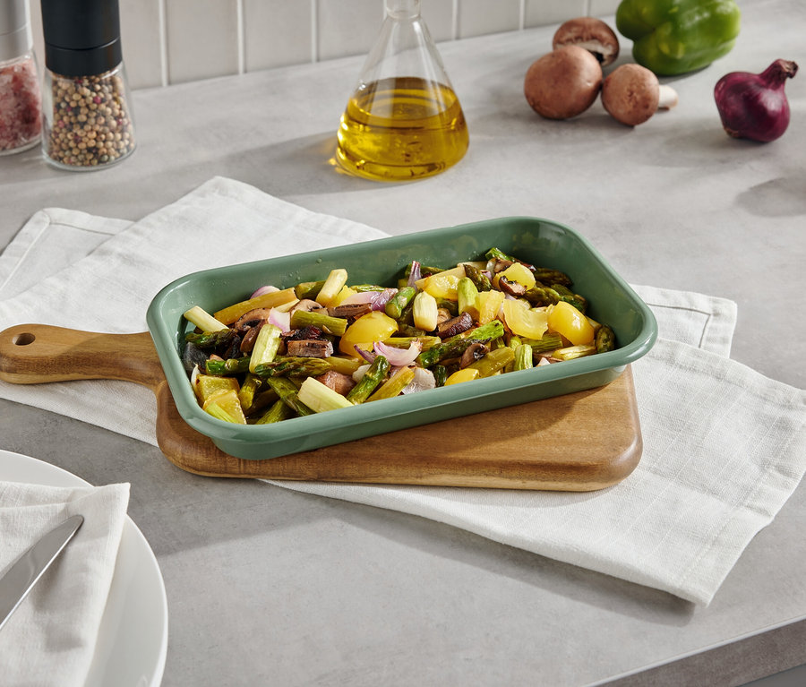 Zelená zapekacia misa s pečenou zeleninou na doske s obrúskom. V pozadí sú huby, paprika, cibuľa a koreniny.