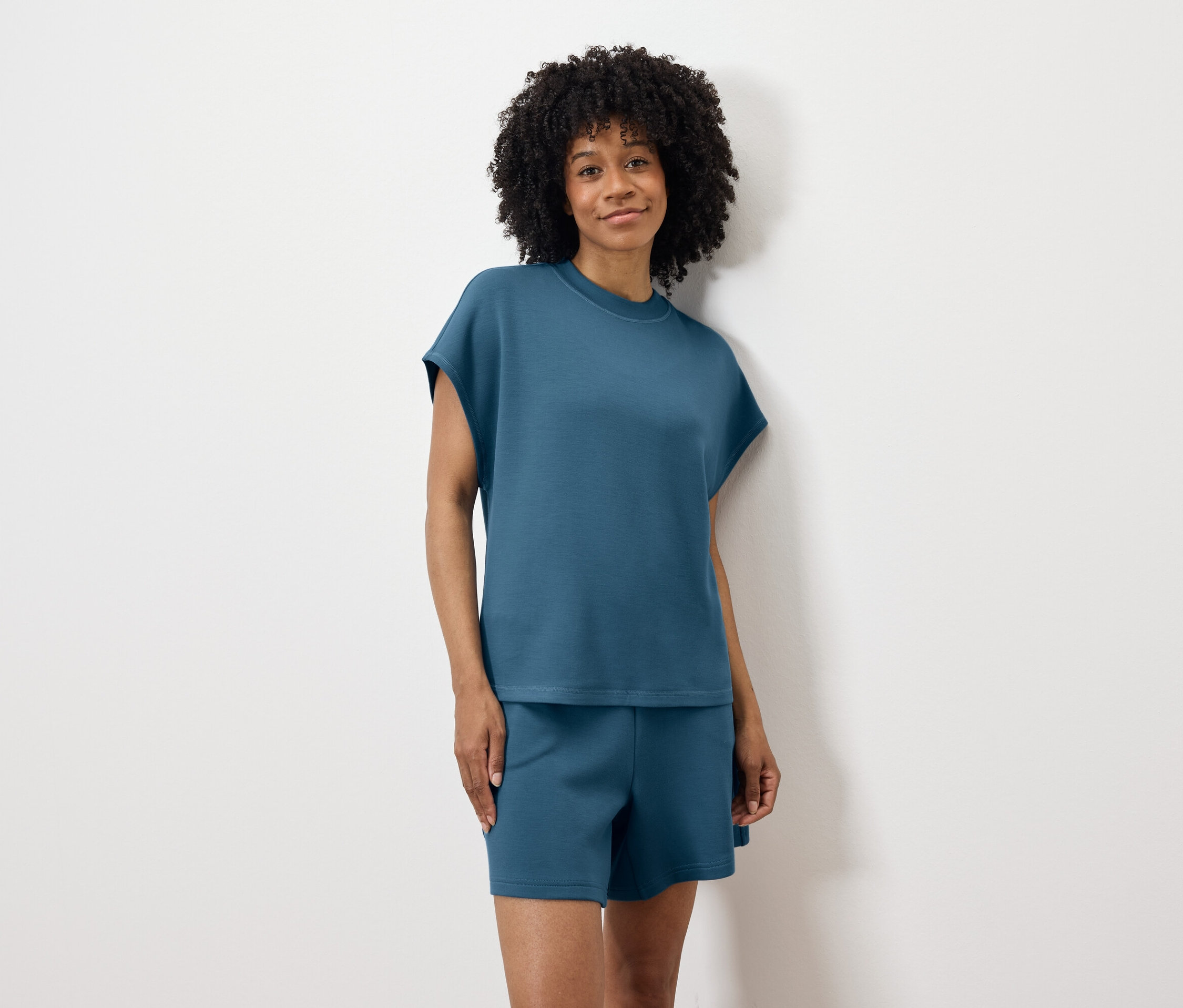 Žena s kučeravými vlasmi v modrom tričku a šortkách stojí pred bielou stenou.