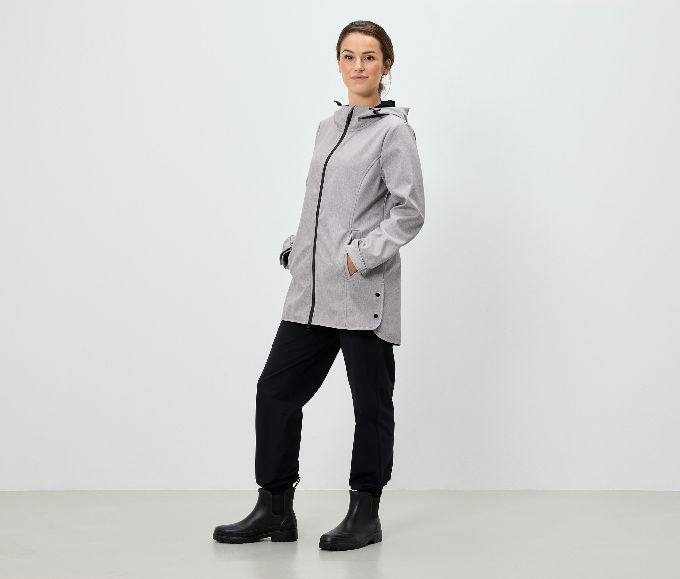Žena stojí a má ruky vo vreckách šedej bundy s kapucňou, čierne nohavice a čierne topánky.