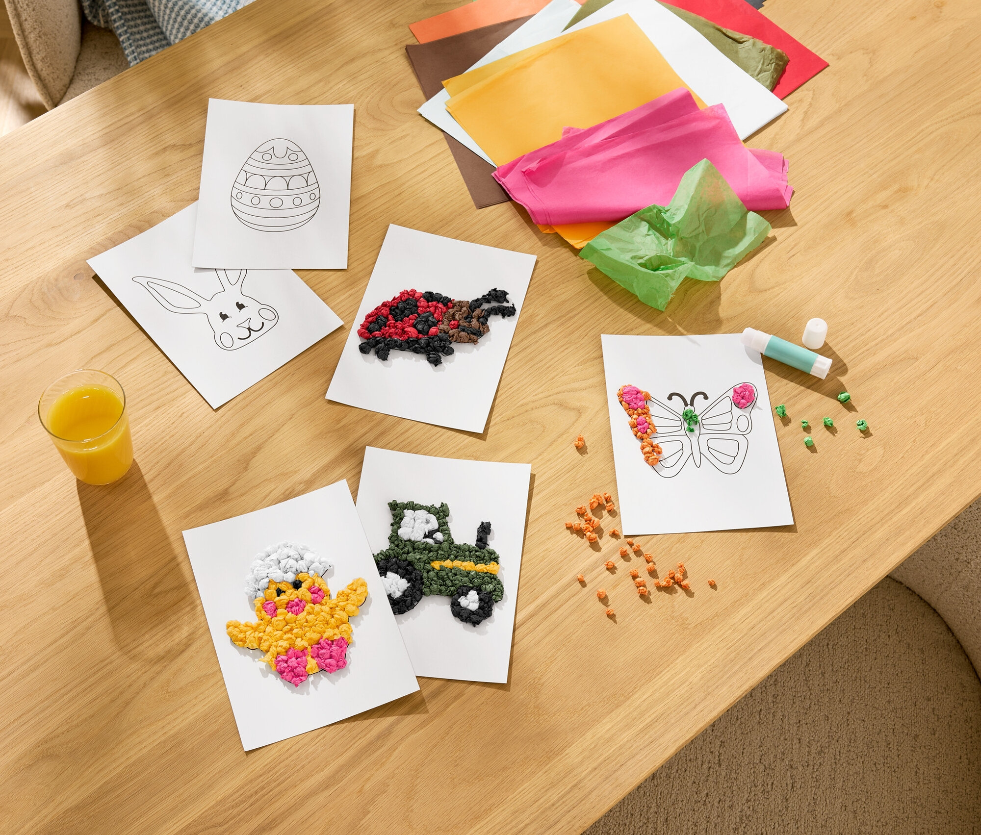 Na drevenom stole ležia ručné práce vrátane nalepených kartičiek s kuriatkom, traktorom, motýľom a korytnačkou. Vidieť aj farebný hodvábny papier, tyčinkové lepidlo a pohár džúsu.