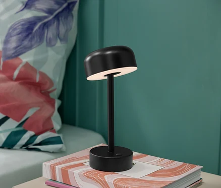 Čierna akumulátorová stolová LED lampa na knihe s kvetinovým vankúšom v pozadí.