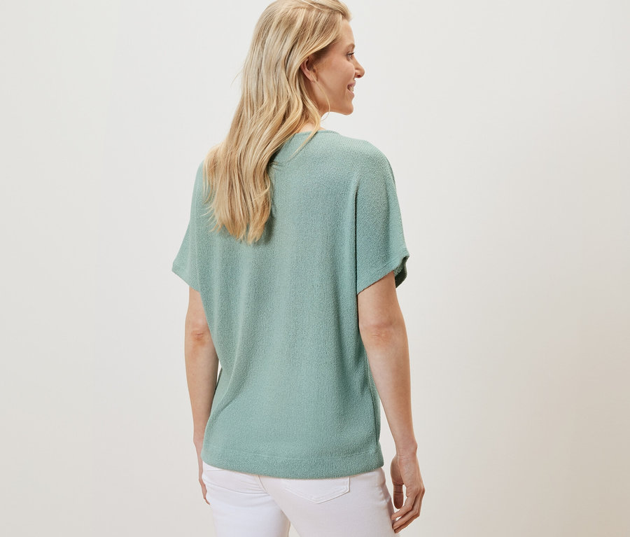 Pohľad zozadu na blondínku v štruktúrovanom tričku šalviovej farby a bielych nohaviciach.