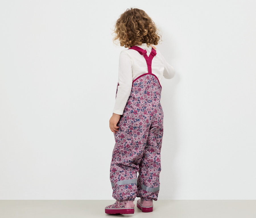 Dievča v kvetinových nohaviciach s náprsenkou a ružových topánkach stojí chrbtom k divákovi.