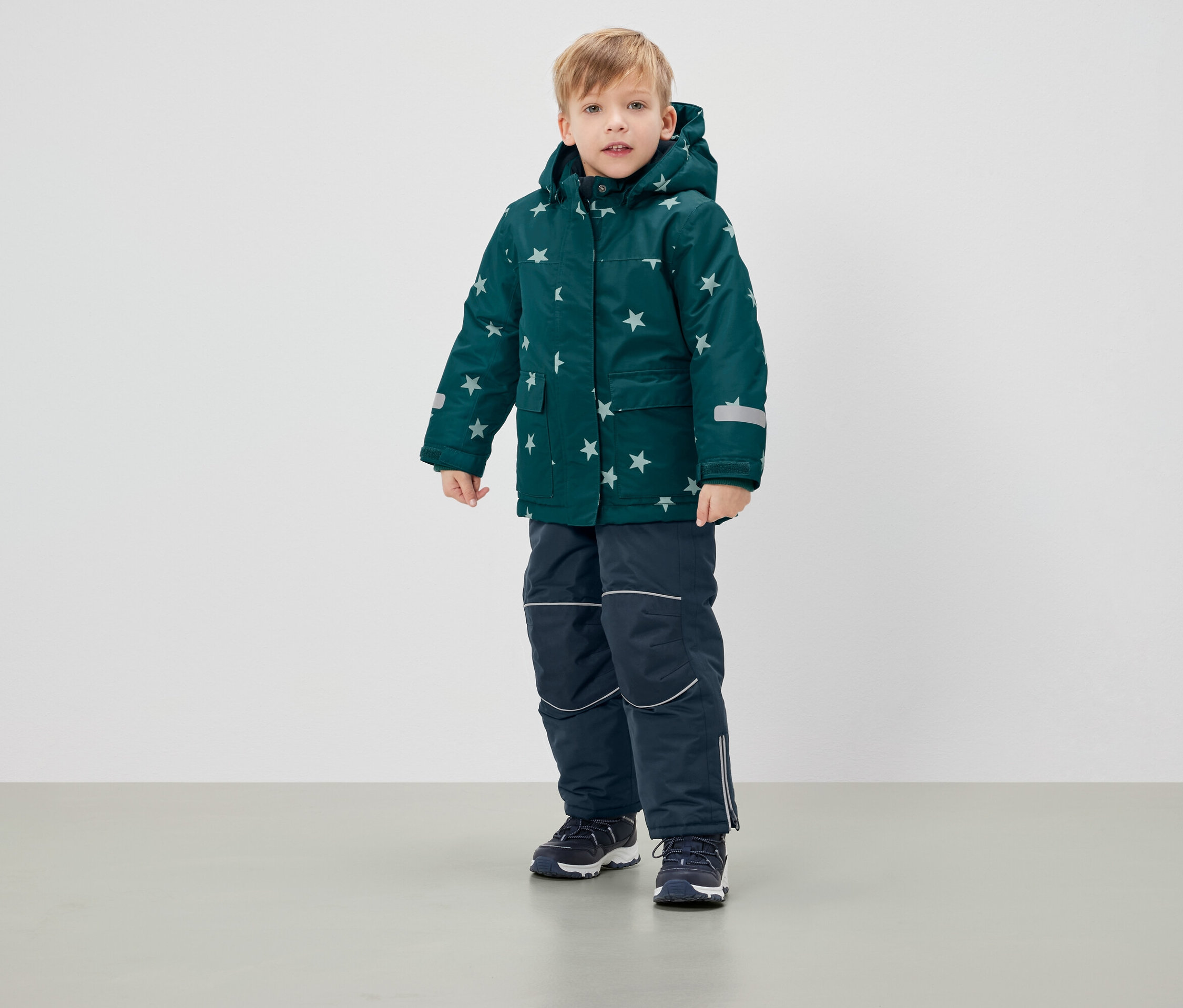 Chlapec stojí v zelenej zimnej bunde s hviezdami a tmavomodrých nohaviciach.