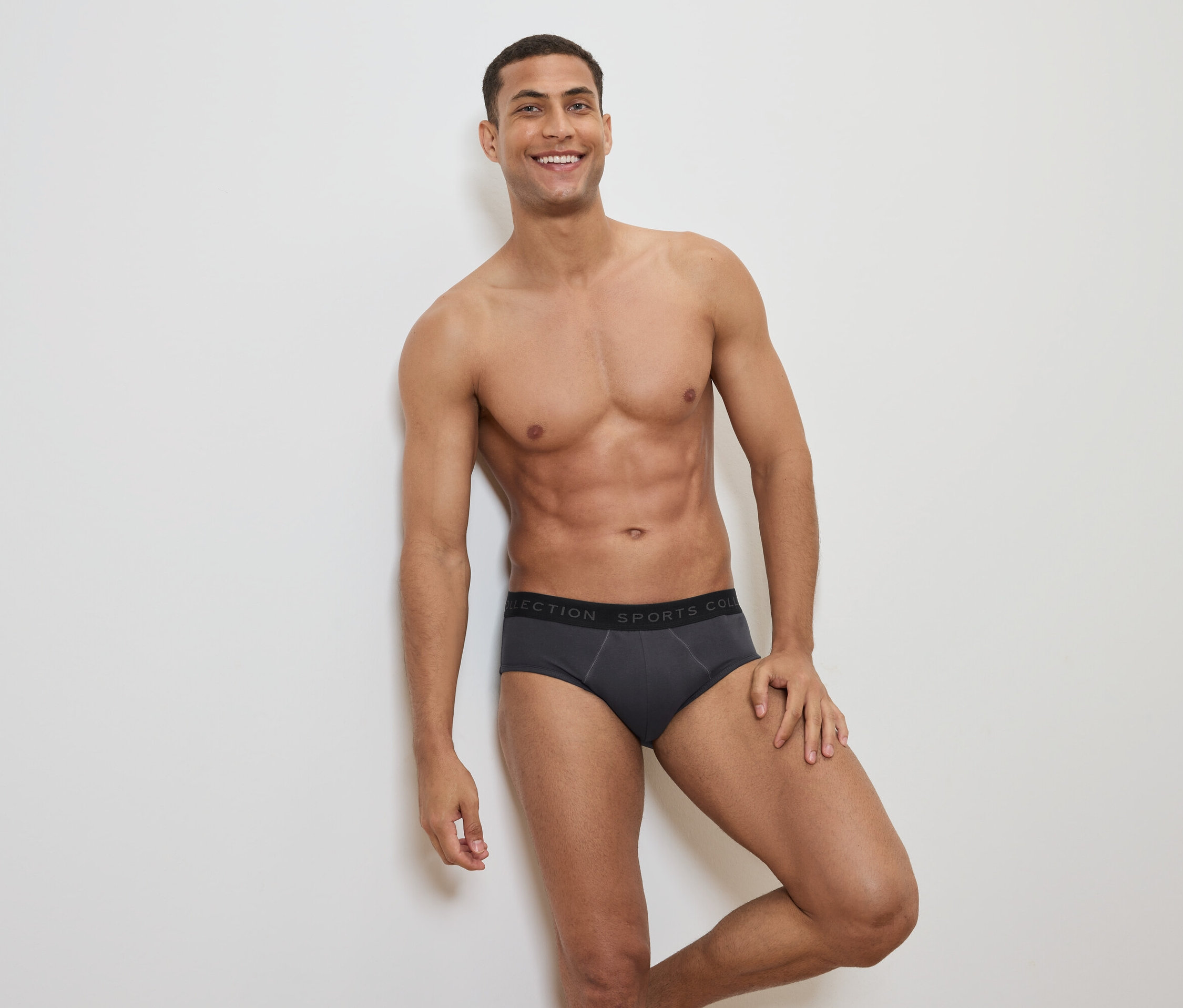 Usmievajúci sa muž s odhalenou hornou časťou tela pózuje v sivých slipových nohavičkách s čiernym pásom.