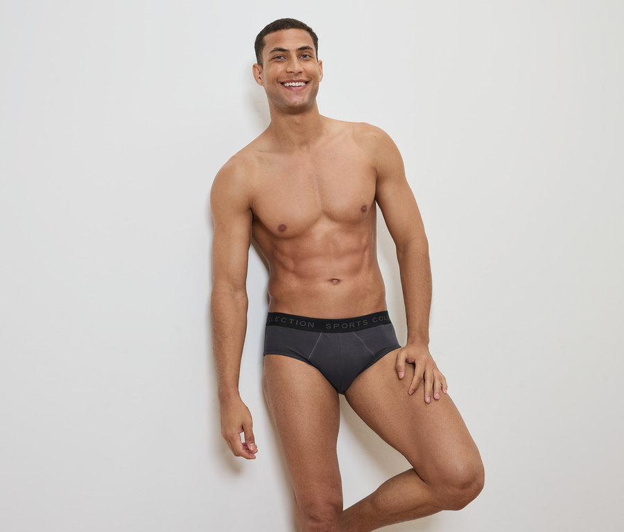 Usmievajúci sa muž s odhalenou hornou časťou tela pózuje v sivých slipových nohavičkách s čiernym pásom.