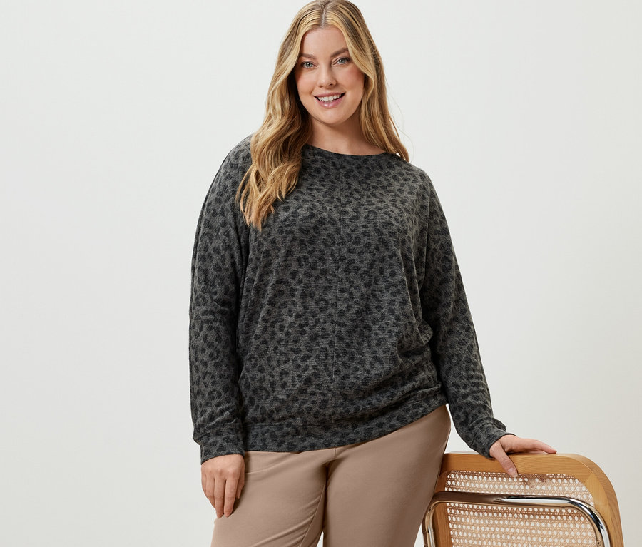 Žena pózuje v béžových teplákových nohaviciach a tričku s netopierími rukávmi s potlačou.