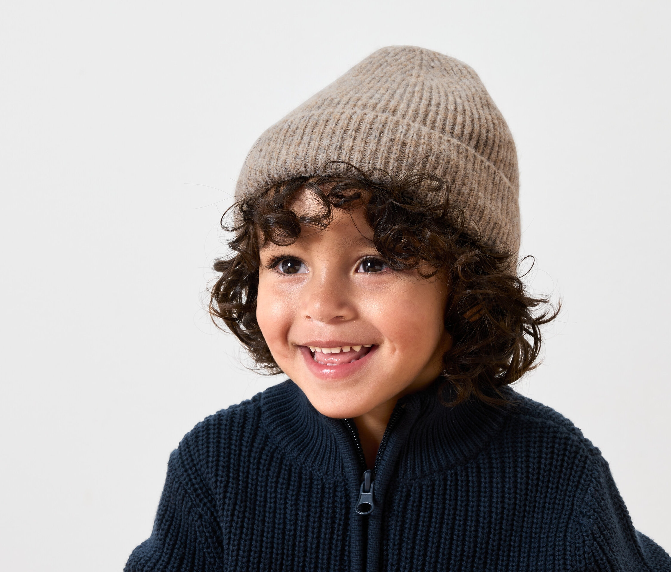 Usmievajúci sa chlapec s hnedými kučeravými vlasmi má na sebe béžovú vlnenú čiapku a tmavomodrý pletený sveter na zips.