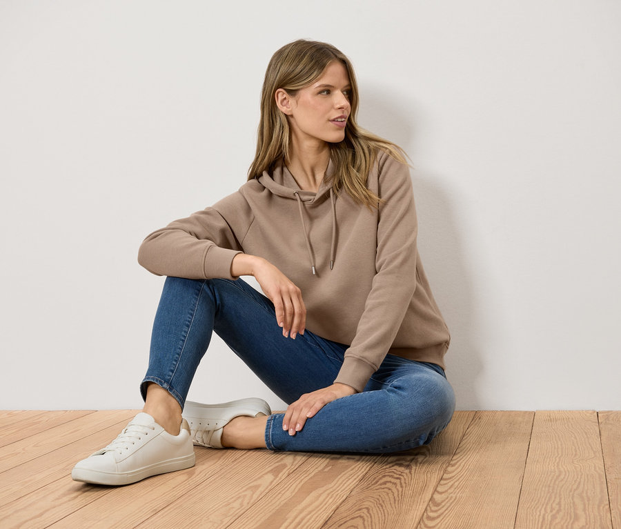 Žena sedí na drevenej podlahe, oblečená v hnedej mikine s kapucňou, modrých džínsoch a bielych teniskách.