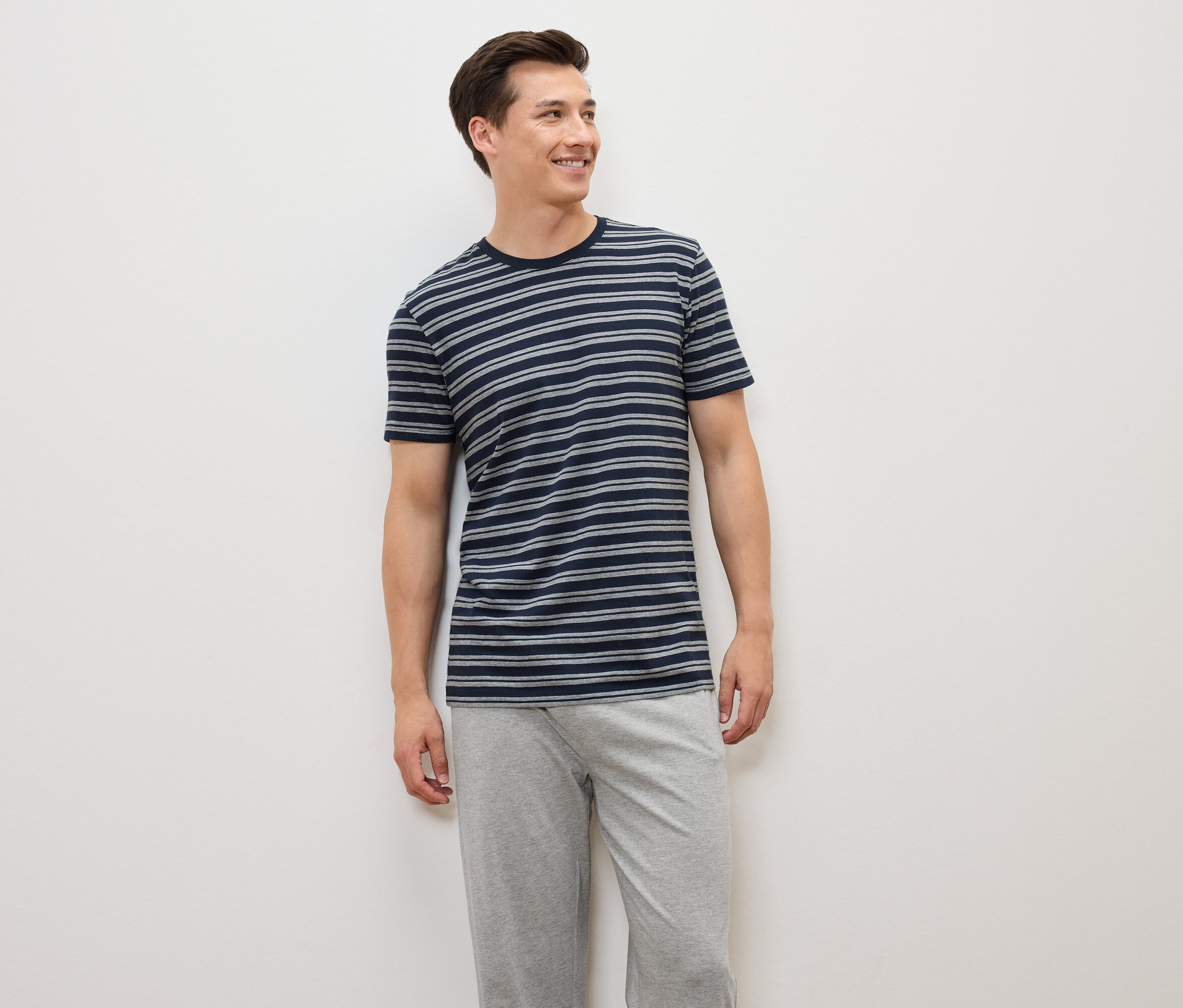 Muž stojí a má na sebe modro-sivé pruhované tričko a sivé nohavice.