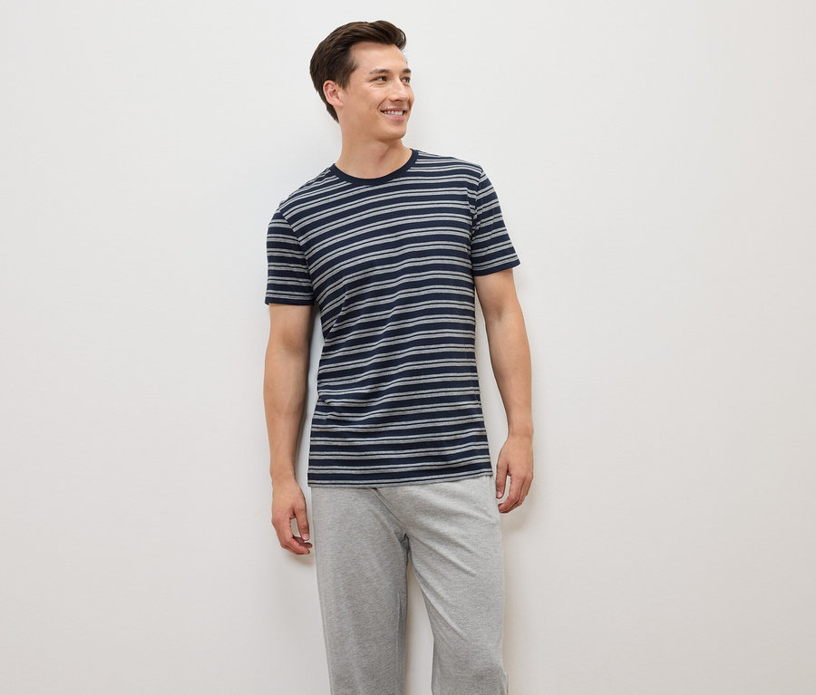 Muž stojí a má na sebe modro-sivé pruhované tričko a sivé nohavice.