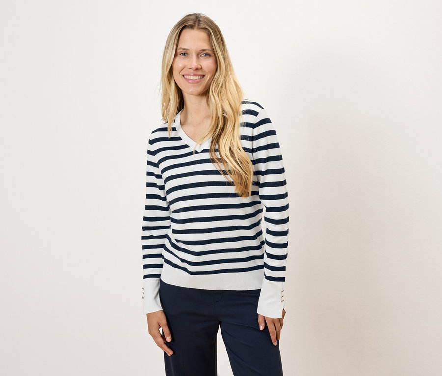 Žena s dlhými blond vlasmi má na sebe pruhovaný sveter a tmavomodré nohavice.