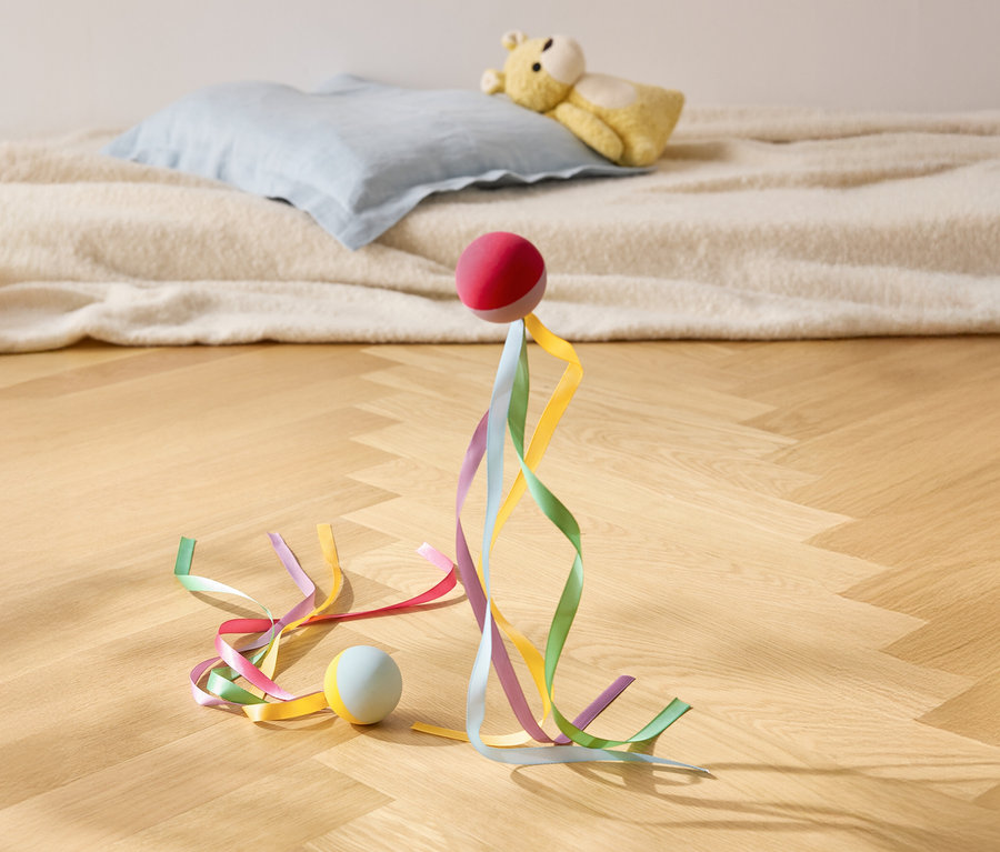 Červená lopta stojí s farebnými stuhami na drevenej podlahe, v pozadí vankúš a plyšový medvedík.