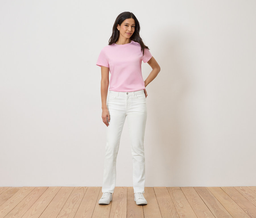 Žena stojí pred bielou stenou a má na sebe ružové tričko, biele džínsy a biele tenisky.