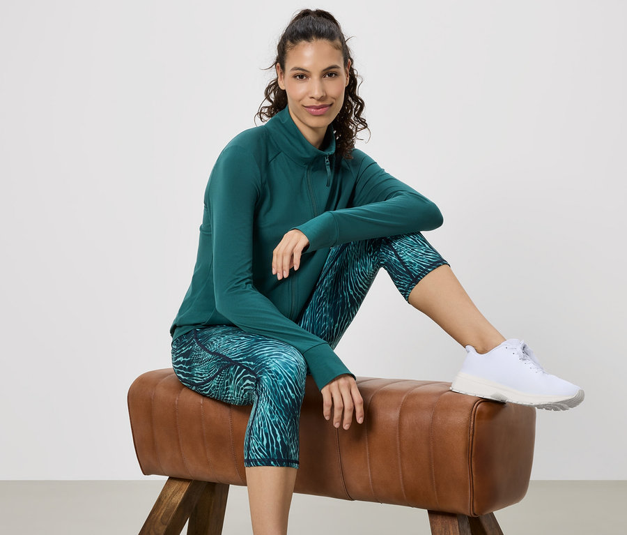 Žena sedí na gymnastickom koni, má na sebe zelenú športovú bundu, vzorované legíny a biele tenisky.