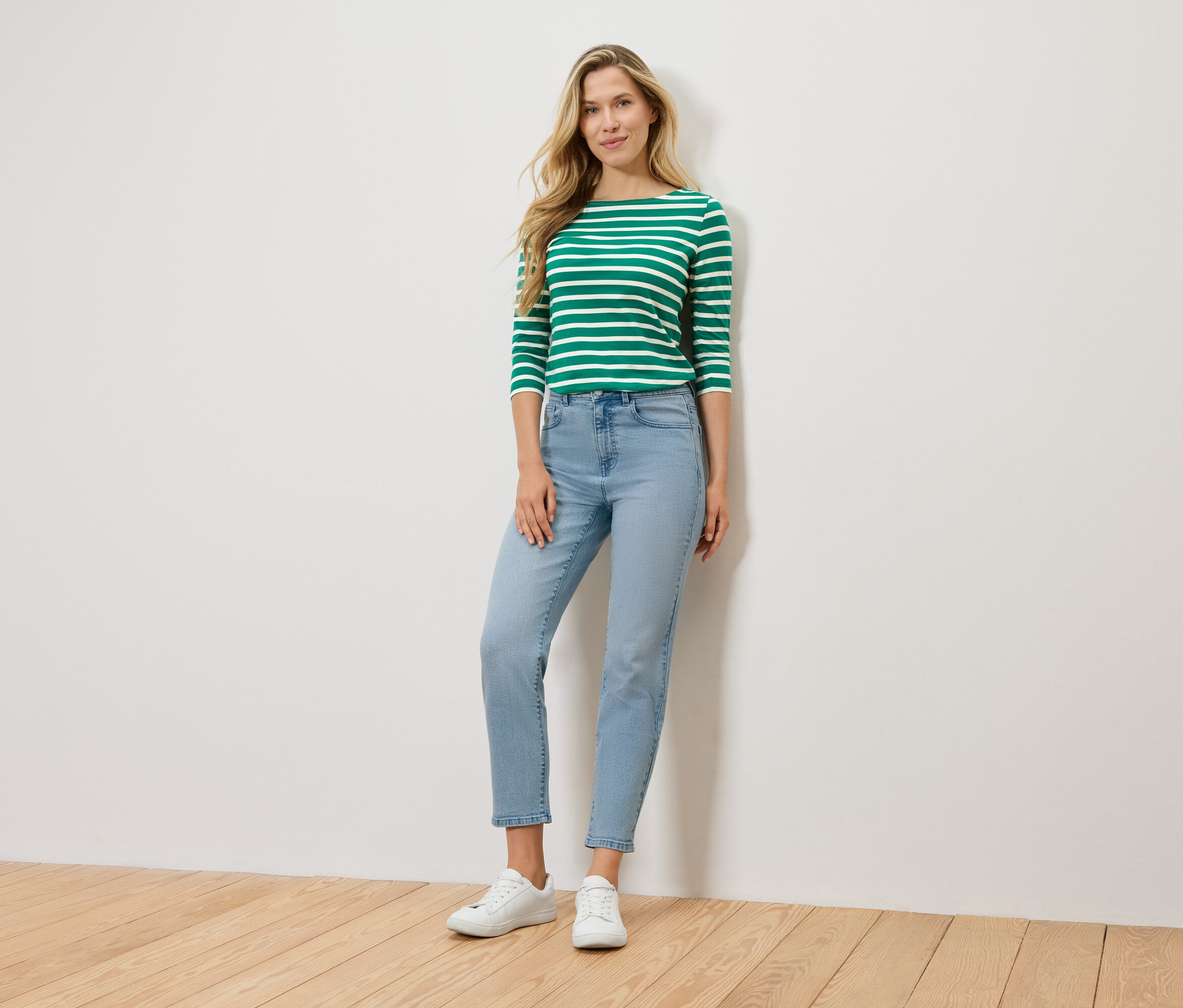 Blondínka stojí pred stenou a má na sebe zeleno-biele pruhované tričko s dlhým rukávom, svetlomodré rifle a biele tenisky.