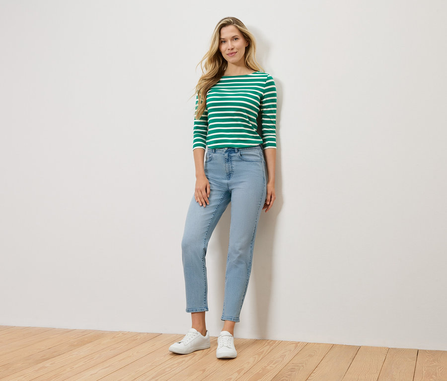 Blondínka stojí pred stenou a má na sebe zeleno-biele pruhované tričko s dlhým rukávom, svetlomodré rifle a biele tenisky.