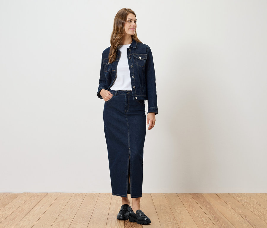 Žena stojí a má na sebe džínsovú bundu a džínsovú sukňu s rozparkom.