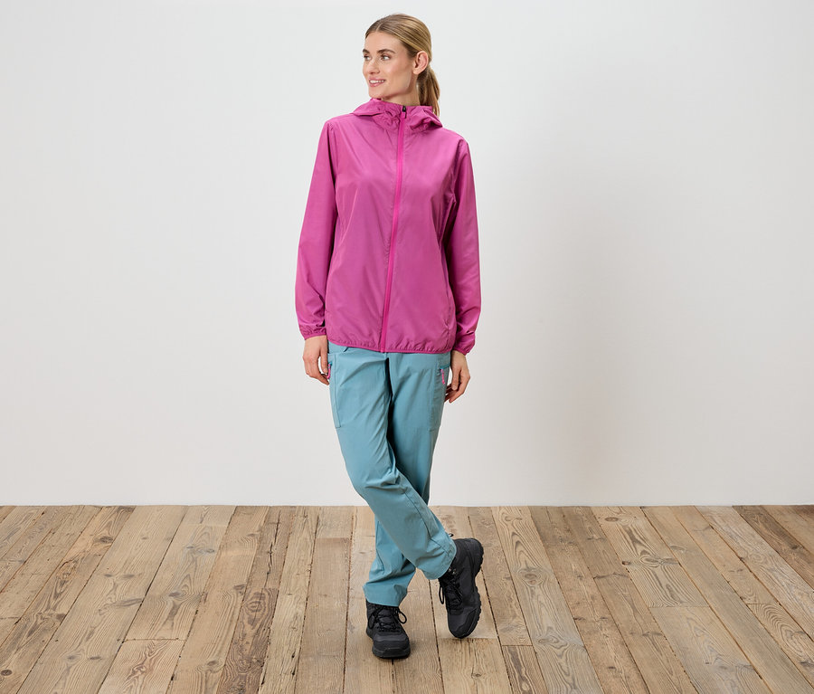 Žena stojí so skríženými nohami, má na sebe ružovú bundu, modré nohavice a čierne topánky.