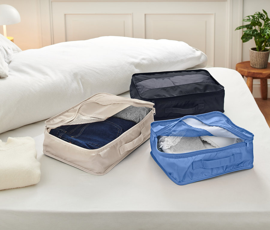 Na posteli s bielymi obliečkami ležia tri otvorené baliace kocky. Kocky sú béžové, modré a čierne a obsahujú oblečenie.