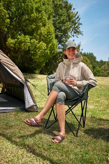 Žena sedí v kempingovej stoličke Tambu »ASANA«, drží šálku a má na sebe 3/4 outdoorové legíny a mikinu s kapucňou.