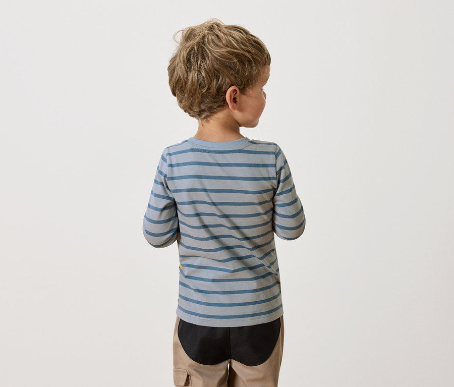 Pohľad zozadu na chlapca s blond kučeravými vlasmi, oblečeného v modro-sivom pruhovanom tričku s dlhým rukávom a béžových nohaviciach.