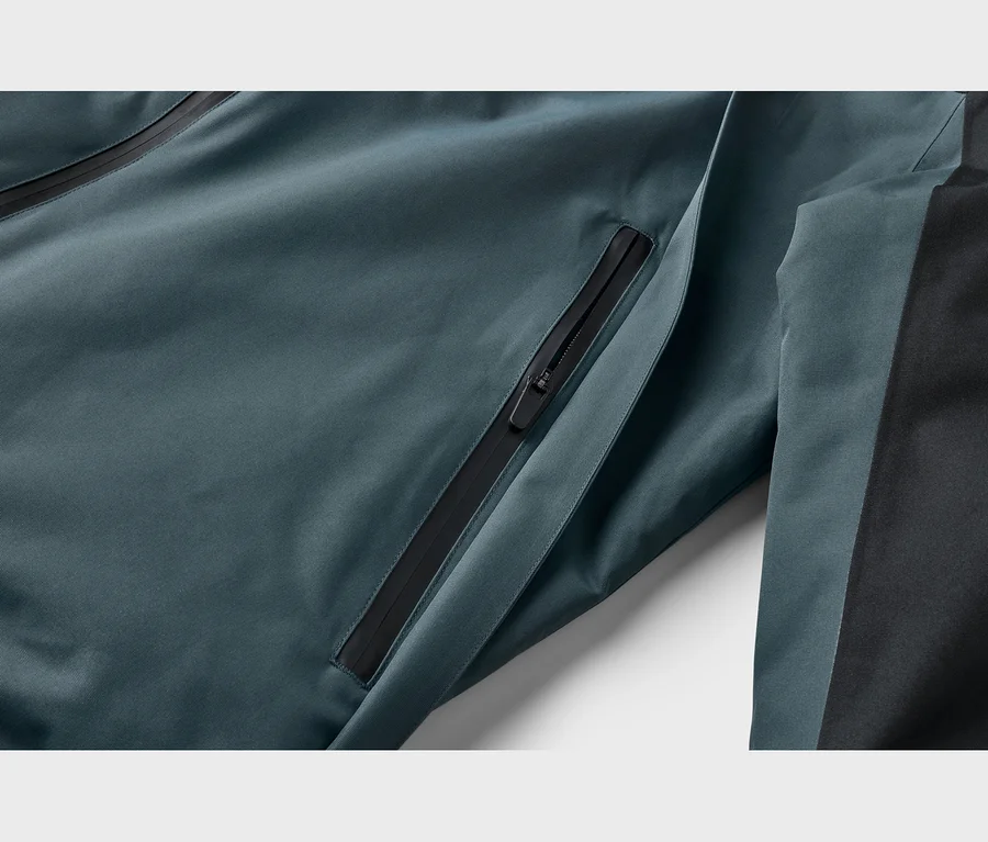 Detail tyrkysovej bundy s čiernym vreckom na zips a čiernym bočným panelom.