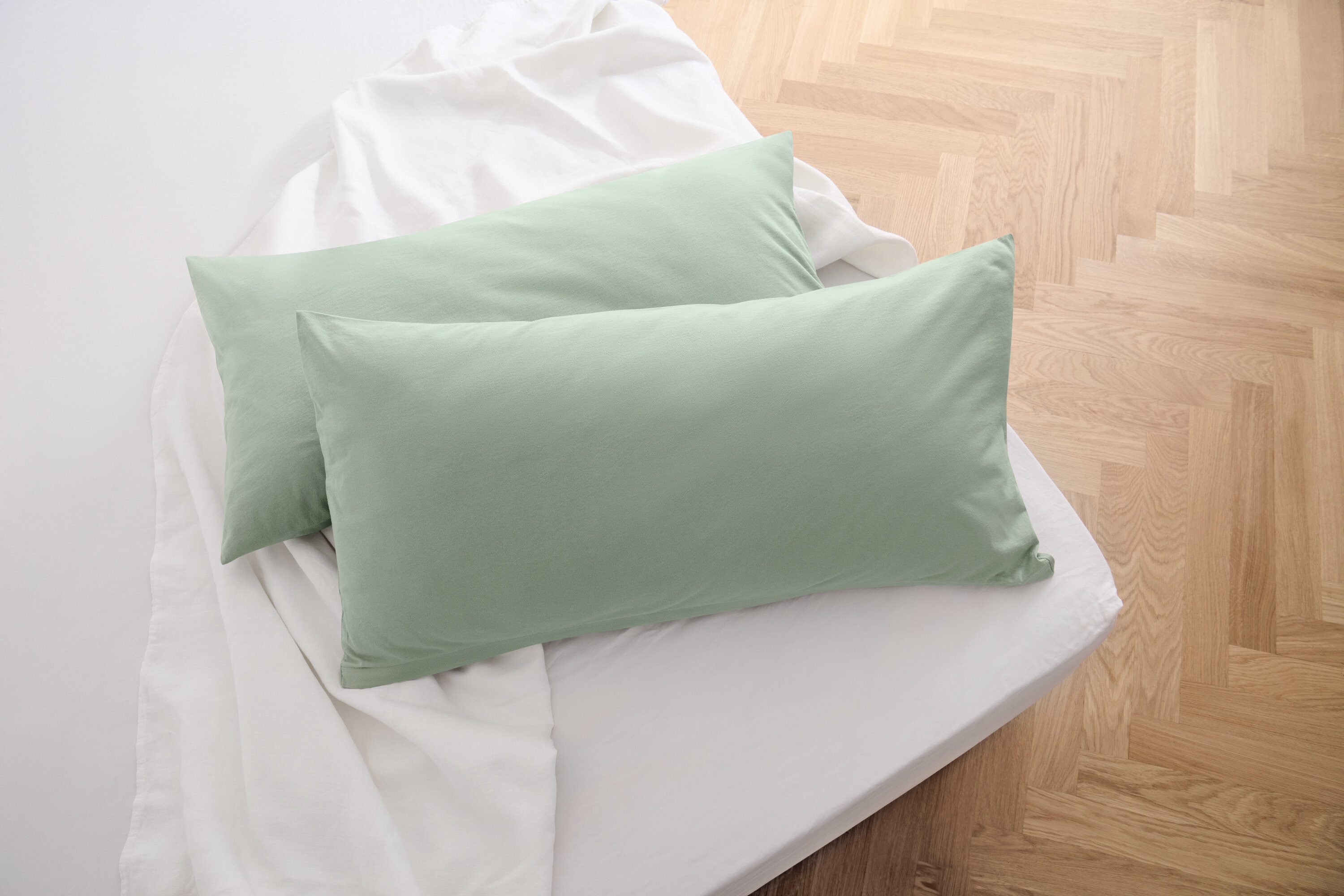 Dve džersejové obliečky na vankúše šalviovozelenej farby ležia na bielej plachte.