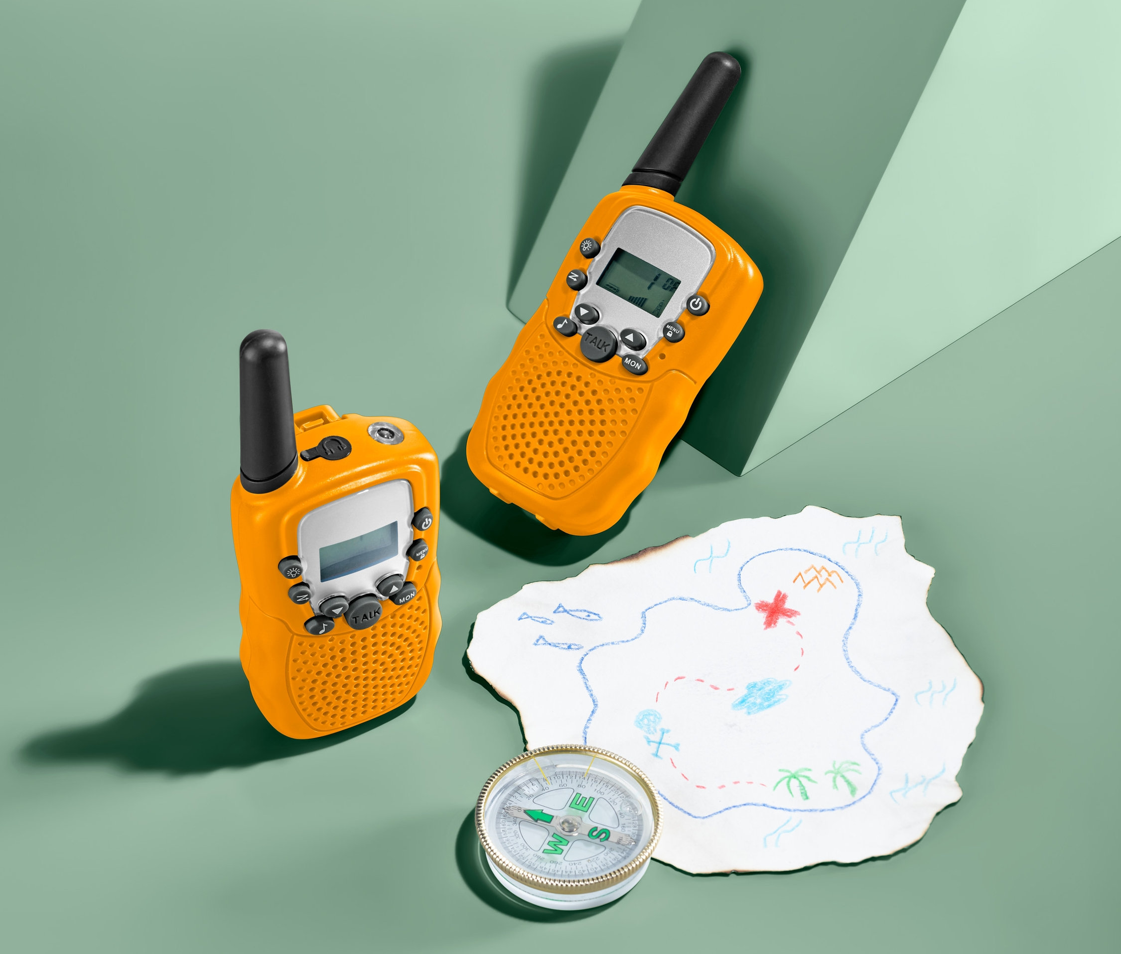 Dve žlté vysielačky a kompas ležia vedľa mapy pokladu na zelenom pozadí.