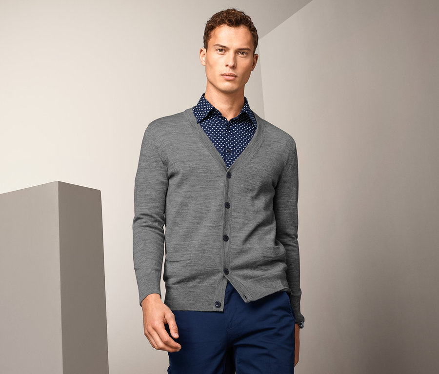 Muž má na sebe sivý merino kardigán cez košeľu s bodkami a modré nohavice.