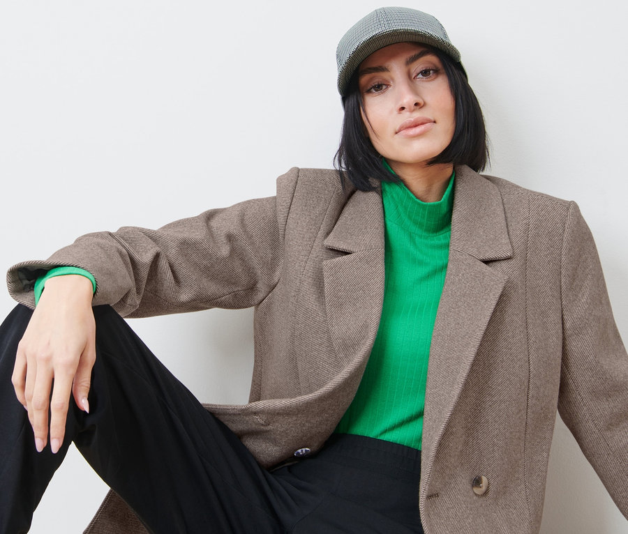 Žena sedí a má na sebe sako, zelený rolák a šiltovku.