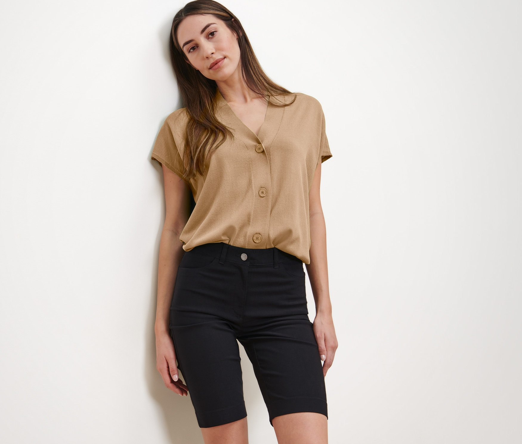 Žena pózuje v béžovej blúzke a čiernych bermudách.